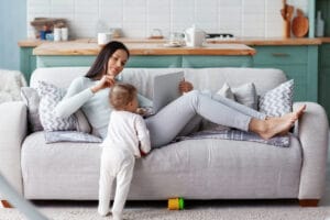 mom works at a laptop at home while her child is playing.