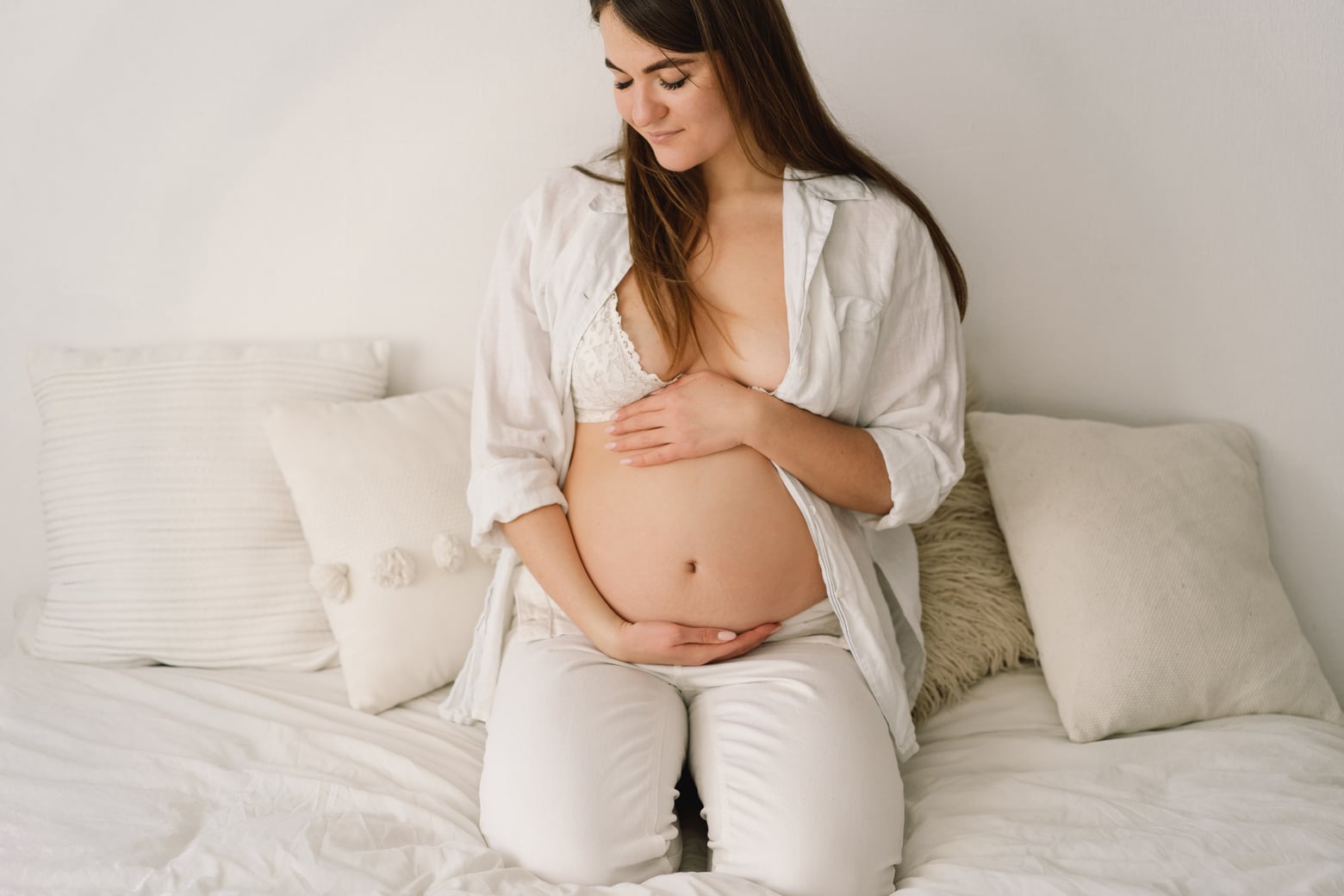 a beautiful pregnant woman, dressed in white clothes, is sitting in bed and holding her belly